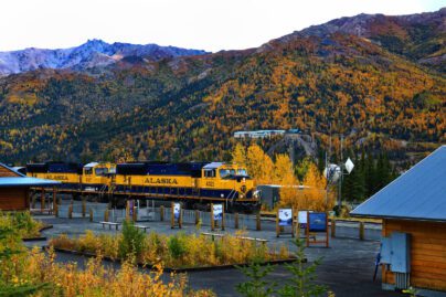 a blue and yellow 'Alaska' train at a depot surrounded by trees in fall colors