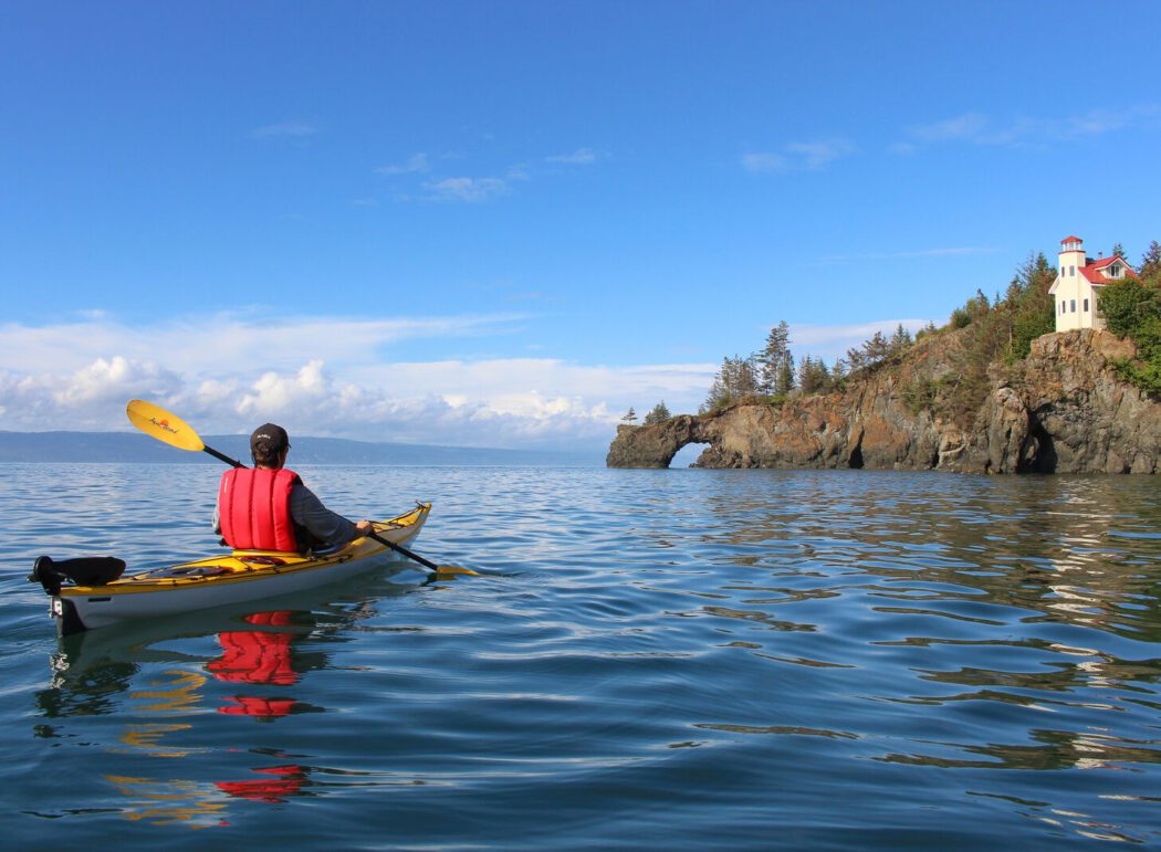 a person in a kayak paddling near a coast with a lighthouse in Kachemak Bay