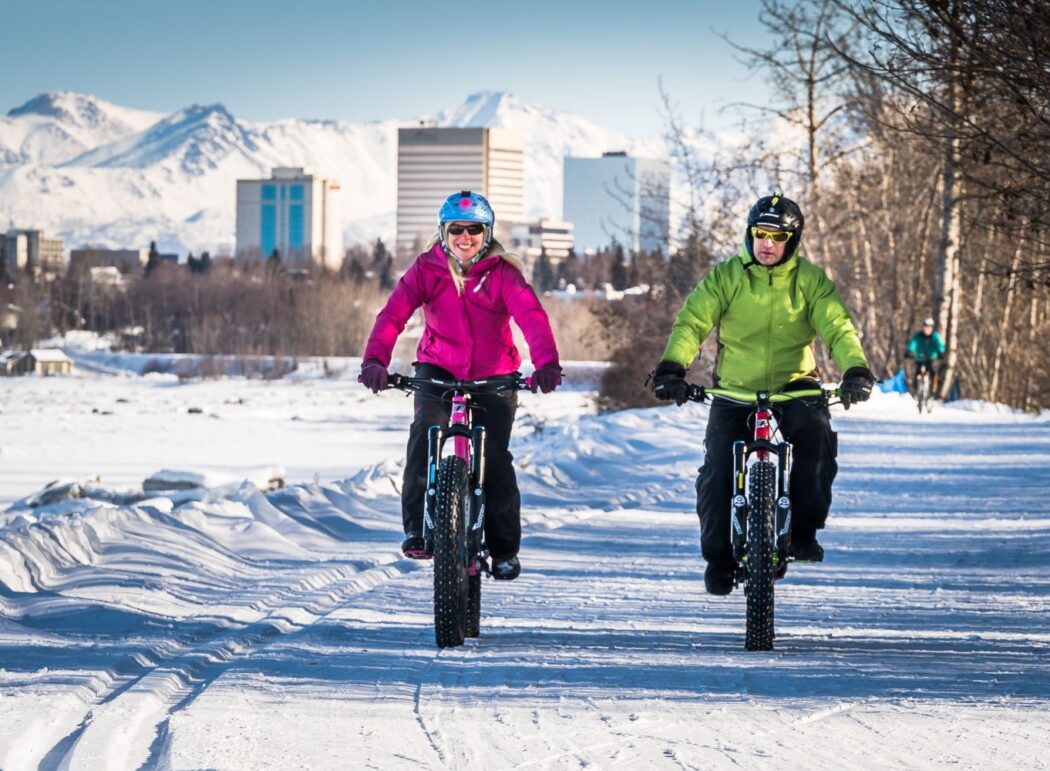 two people on fat tire bikes on a winter trail; Anchorage, Alaska cityscape in background