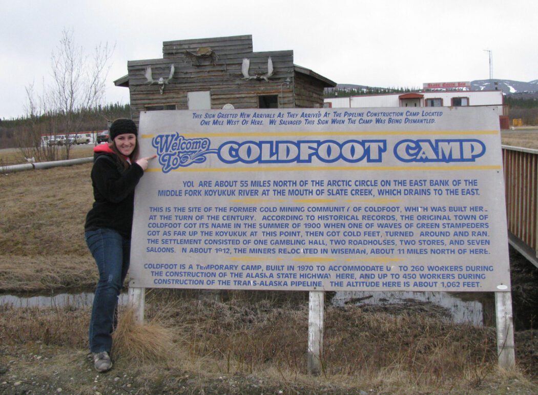 a person pointing at a sign that says "Welcome to Coldfoot Camp. You are about 55 miles north of the Arctic Circle"
