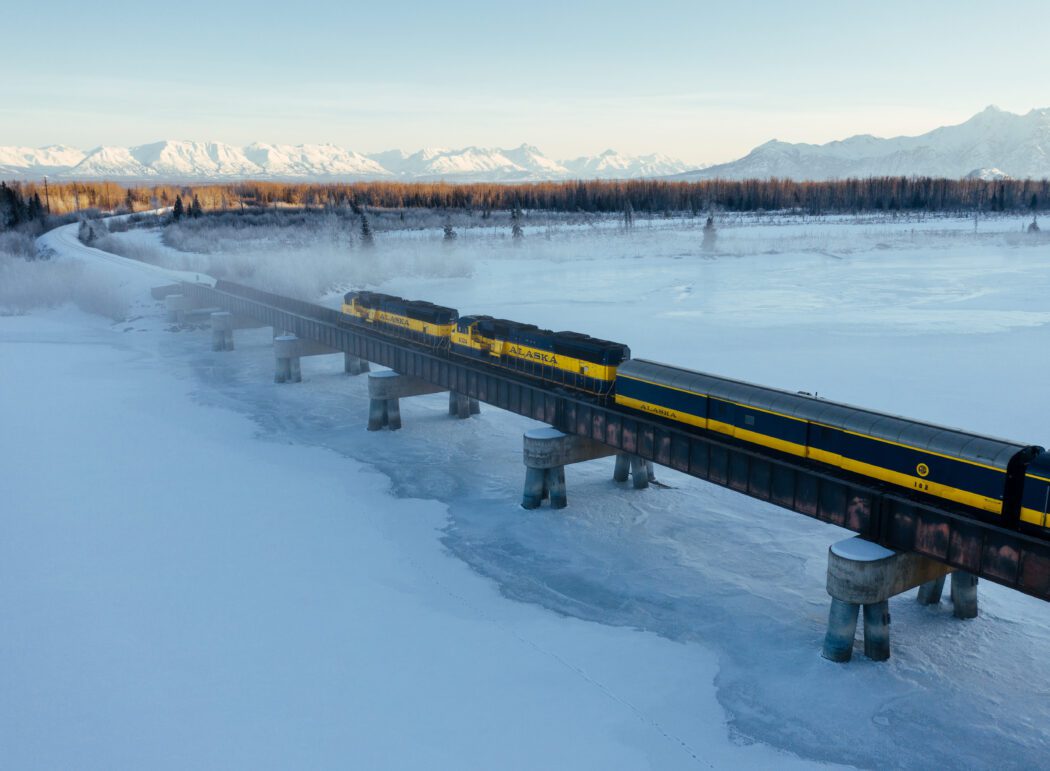a blue and yellow passenger train travels a bridge over a frozen river; snowy mountains in background