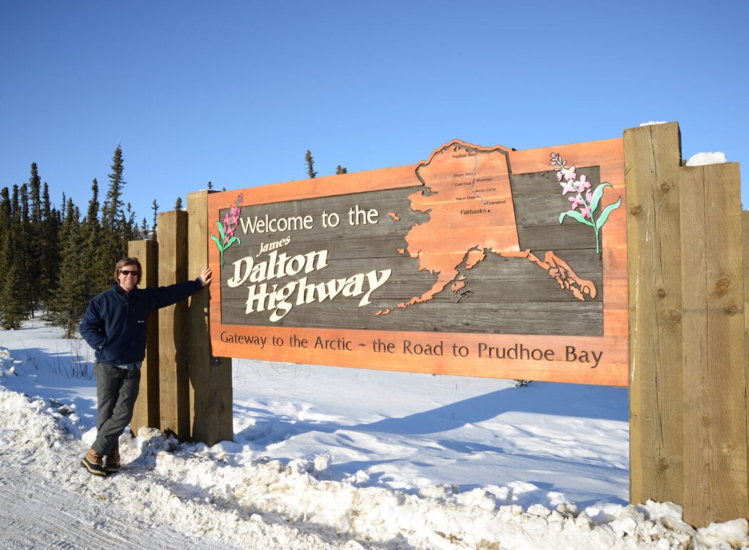 a person on a snowy road leaning on a wooden sign reading "Welcome to the James Dalton Highway, Gateway to the Arctic - Road to Prudhoe Bay "