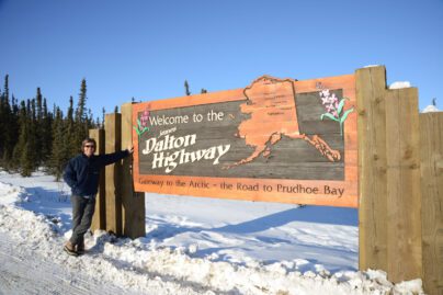 a person on a snowy road leaning on a wooden sign reading "Welcome to the James Dalton Highway, Gateway to the Arctic - Road to Prudhoe Bay "