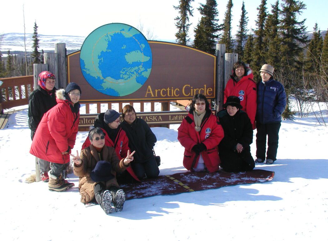 a group of people in winter jackets pose for a photo in front of a wooden sign reading "Arctic Circle, Dalton Highway, Alaska"