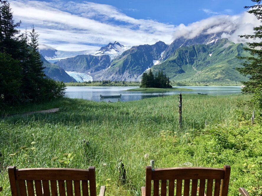 two wooden chairs facing out to grass coast line, a lagoon with a canoe and a tidewater glacier