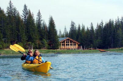two people kayaking in a glacial lagoon in front of an Alaskan wilderness lodge on shore