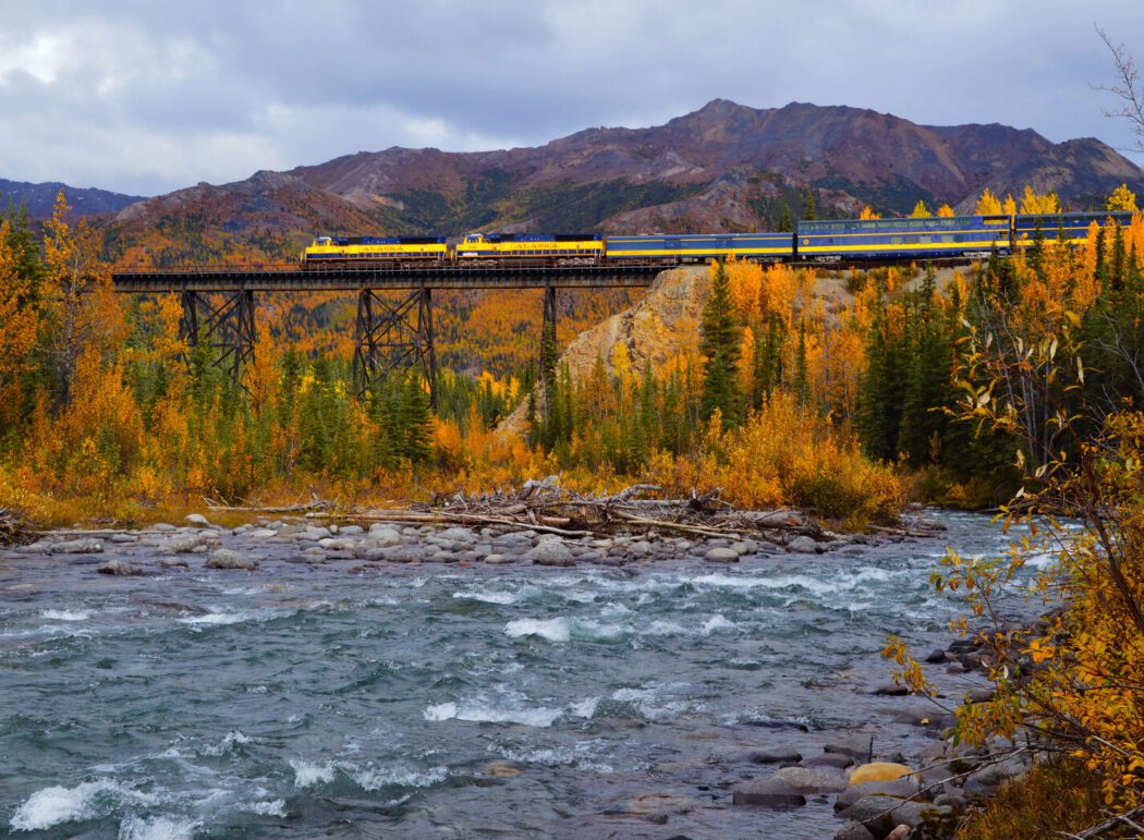 the yellow and blue 'Alaska' passenger train on a bridge over a river in Alaska; mountains and fall foliage around