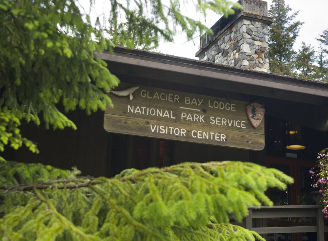 Evergreen trees and a wooden sign on a lodge reading "Glacier Bay Lodge | National Park Service Visitor Center"