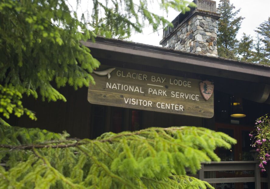 Evergreen trees and a wooden sign on a lodge reading "Glacier Bay Lodge | National Park Service Visitor Center"