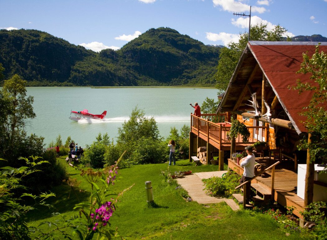 People at a log-cabin lodge watching a float-plane take off from an adjacent lake