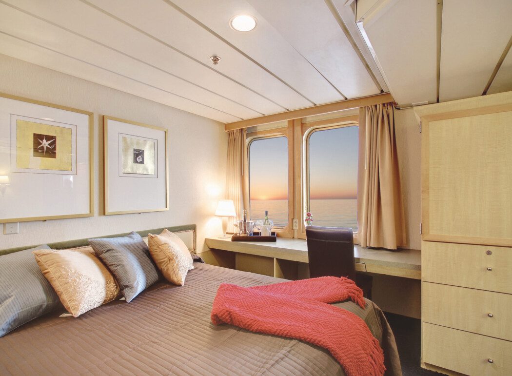 the inside of a cruise ship cabin, showing a bed, a desk with wine on it, and window views to the ocean