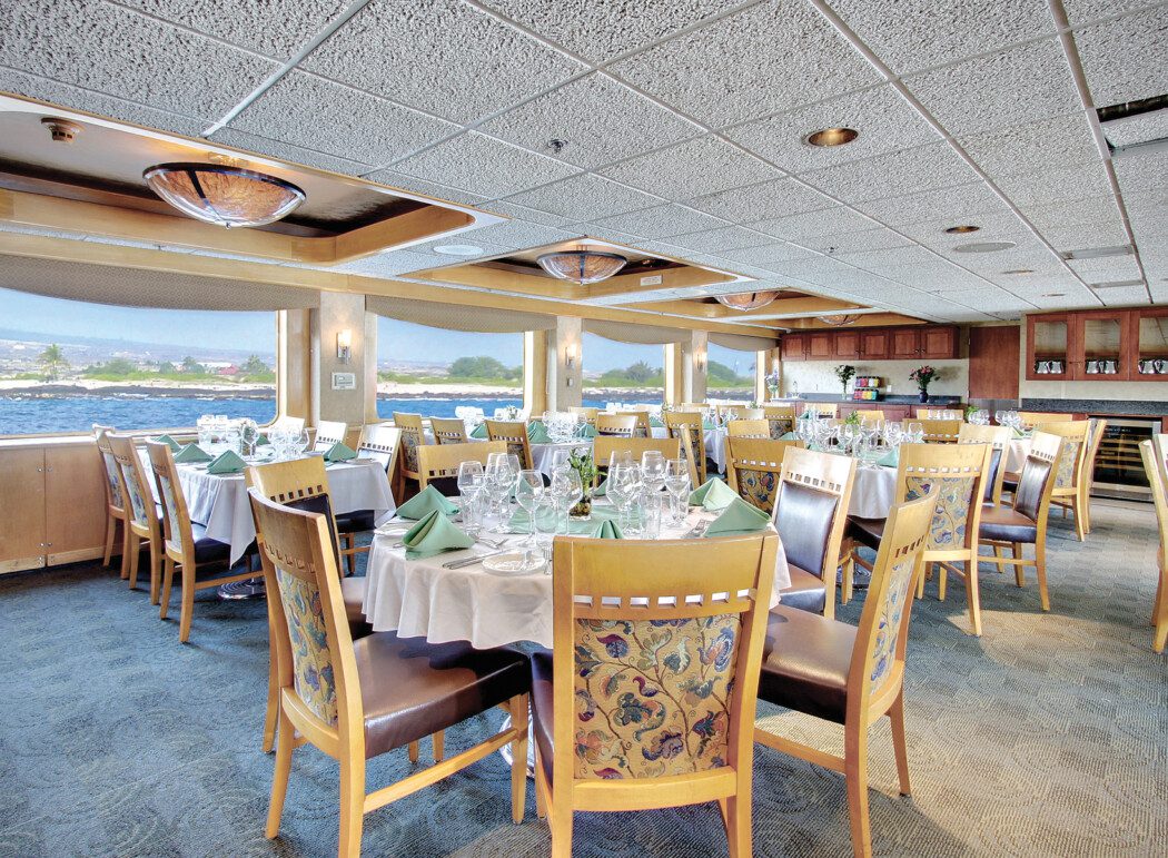 the dining room of a cruise ship, showing tables set, and coastal views from the windows