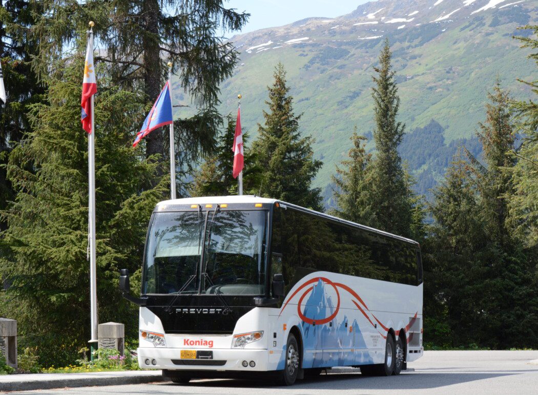 a tour bus parked next to evergreen trees and flag poles; mountains in background