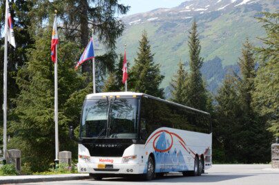 a tour bus parked next to evergreen trees and flag poles; mountains in background