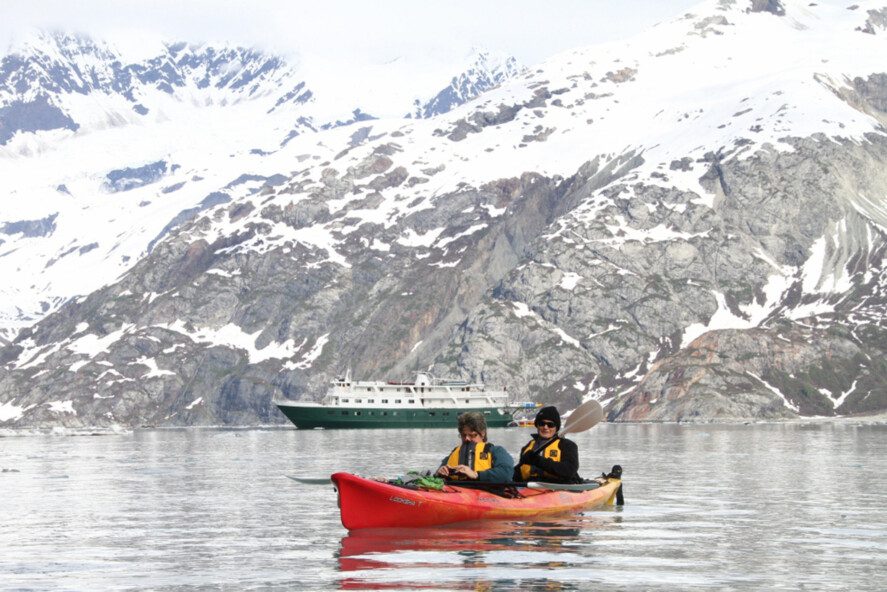 two people in a sea kayak; a small cruise ship and snow-capped mountains in background