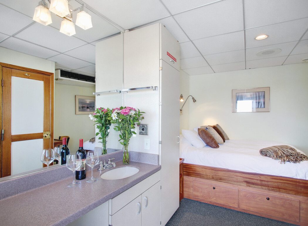 the inside of a cruise ship cabin, showing a bed and a bottle of wine and flowers on a sink