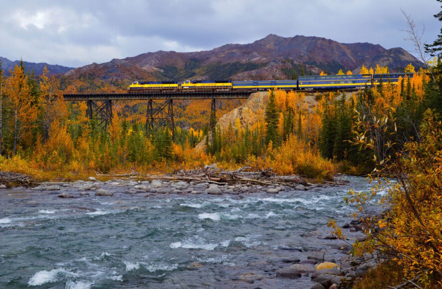 the yellow and blue 'Alaska' passenger train on a bridge over a river in Alaska; mountains and fall foliage around