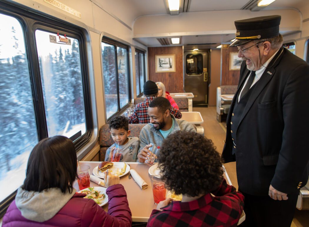 an Alaska Railroad train conductor in a dining car talks to a family enjoying a meal