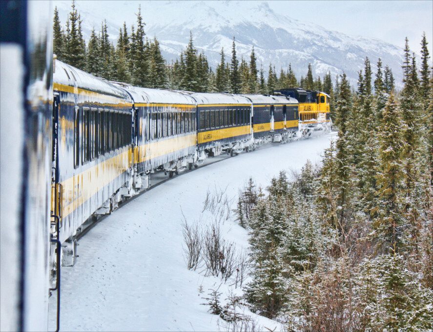a yellow and blue passenger train traveling through a boreal forest in winter