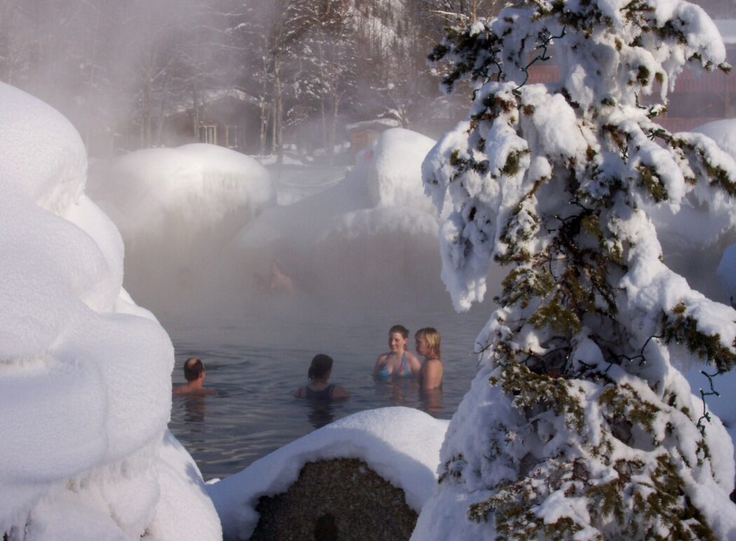 people relax in a hot springs pool surrounded by snow covered rocks and spruce trees