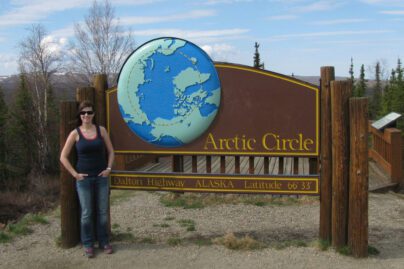 a person standing next to a wooden sign reading "Arctic Circle, Dalton Highway, Alaska, Latitude 66, 33"