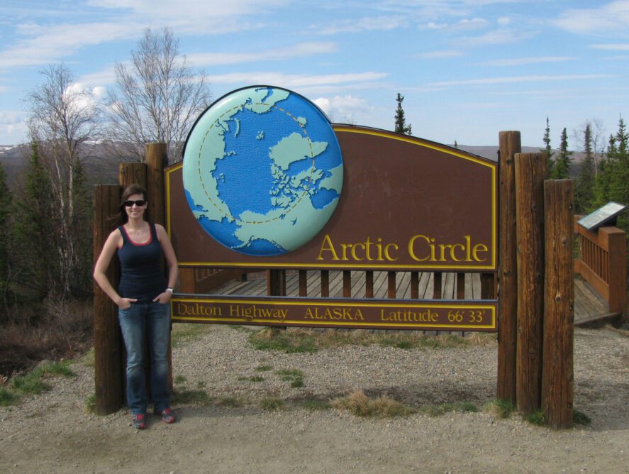 a person standing next to a wooden sign reading "Arctic Circle, Dalton Highway, Alaska, Latitude 66, 33"
