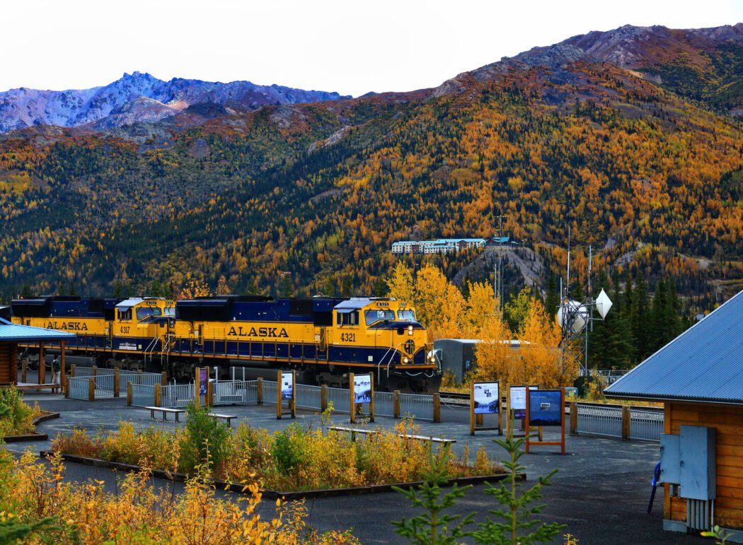 a blue and yellow 'Alaska' train at a depot surrounded by trees in fall colors