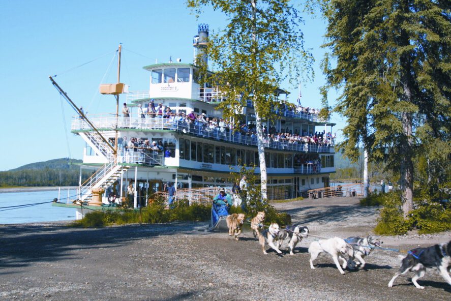 people onboard a sternwheeler along the banks of a river watch a sled dog team pull a sled over gravel in the summertime