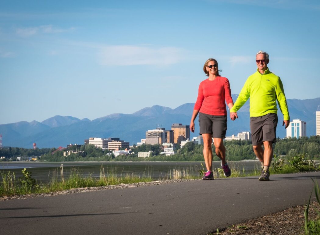 a man and a woman walking on a paved trail; Anchorage, Alaska and mountains in background