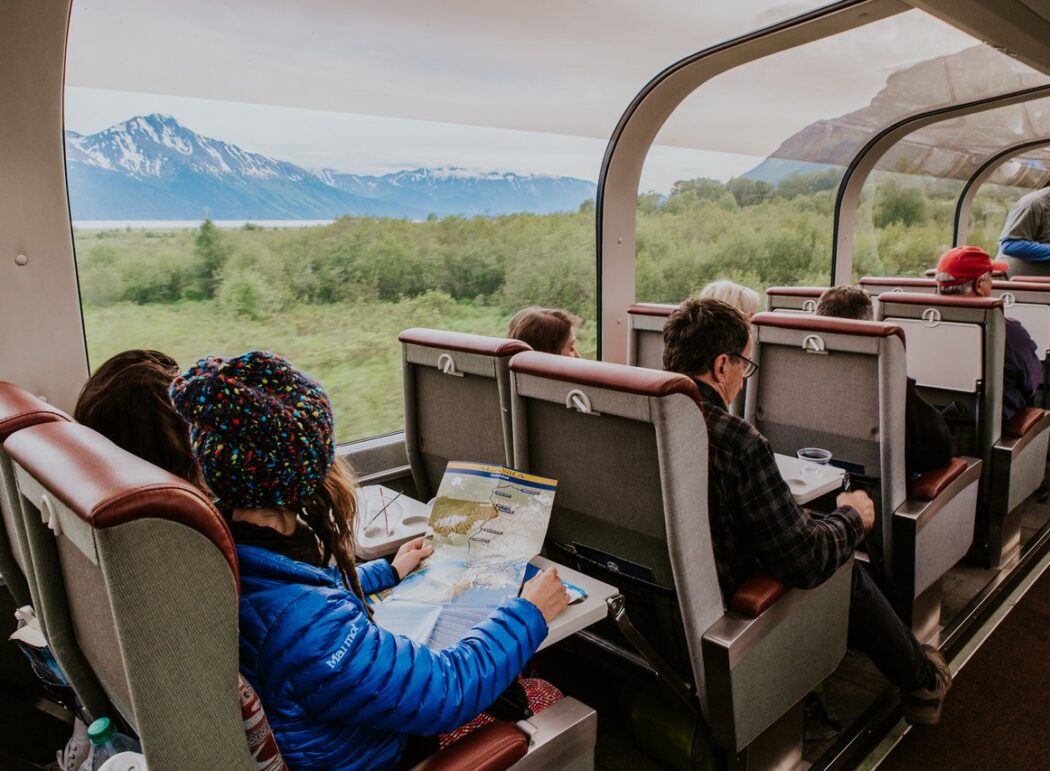 people on a train looking at Alaskan scenery through dome-car windows; one woman holds a map of Alaska