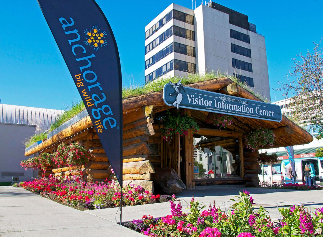 a log cabin visitors center surrounded by flowers. A flag reads "Anchorage: Big Wild Life"