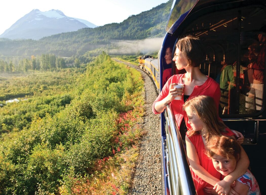 a woman and two children look out at Alaskan scenery from the viewing platform of a passenger train