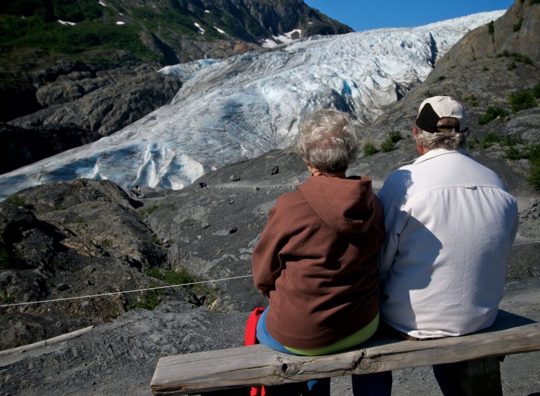 two visitors sit on a park bench with Exit Glacier in front of them