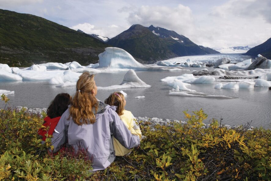 a woman and two children sit on the shore of a glacial lake admiring Spencer Glacier