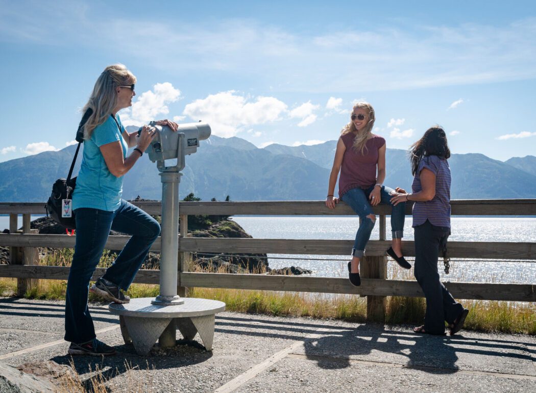 two women sit on a fence while another leans on a pair of mounted binoculars looking out to water and mountains