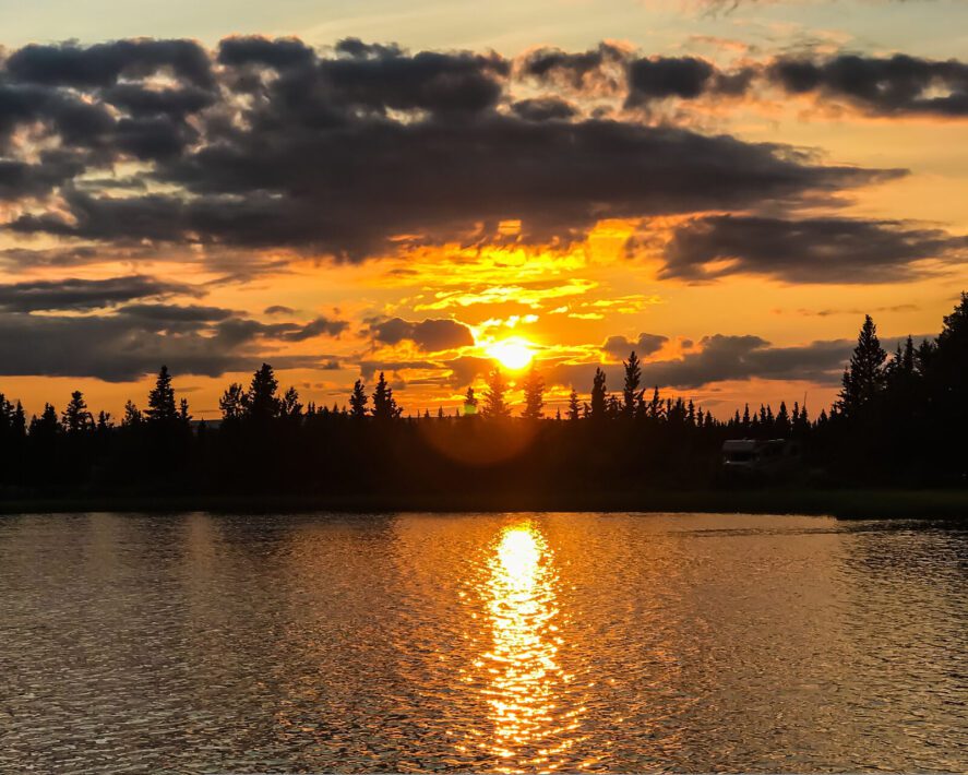 the Midnight Sun hangs above the horizon over spruce trees and water