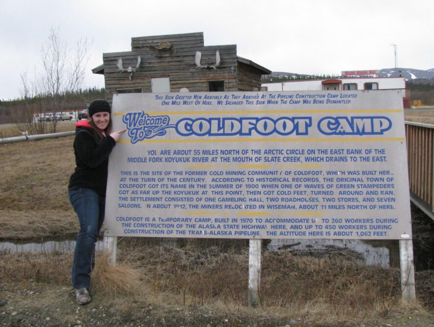 a person standing next to a sign outside a lodge that says "Welcome to Coldfoot Camp"