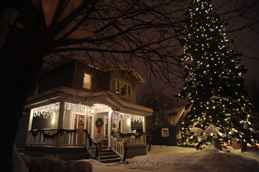 Alaskan house with christmas lights and a huge spruce tree lit up with white lights under a recent snowfall.